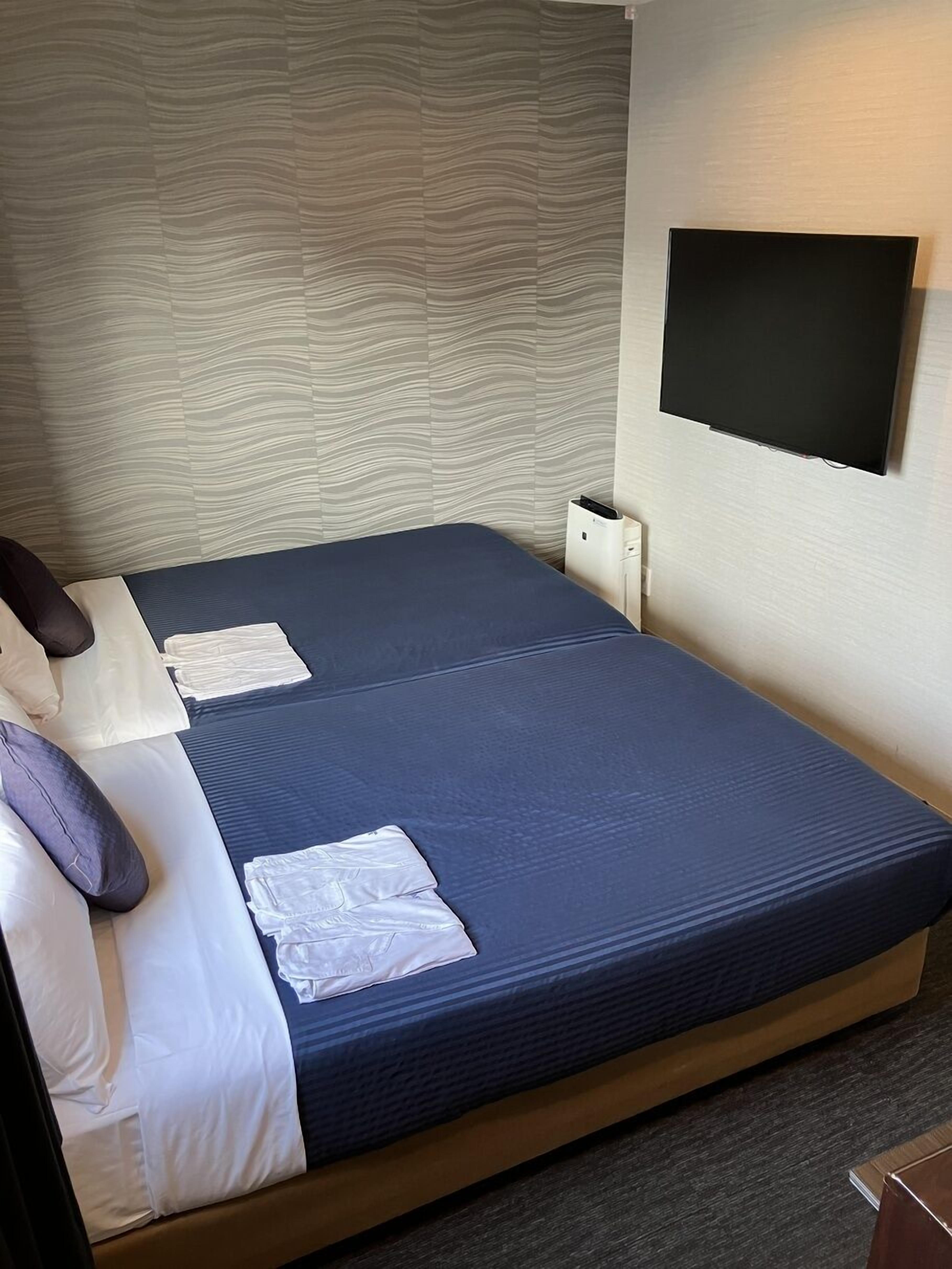 A hotel room with a bed, a flat-screen TV mounted on the wall, and a small bedside table.