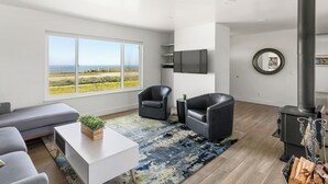 A modern living room with a large window, grey sofa, black leather chairs, a white coffee table, and a wood-burning stove. - Point Arena Lighthouse - Head Keeper's House (Point Arena)