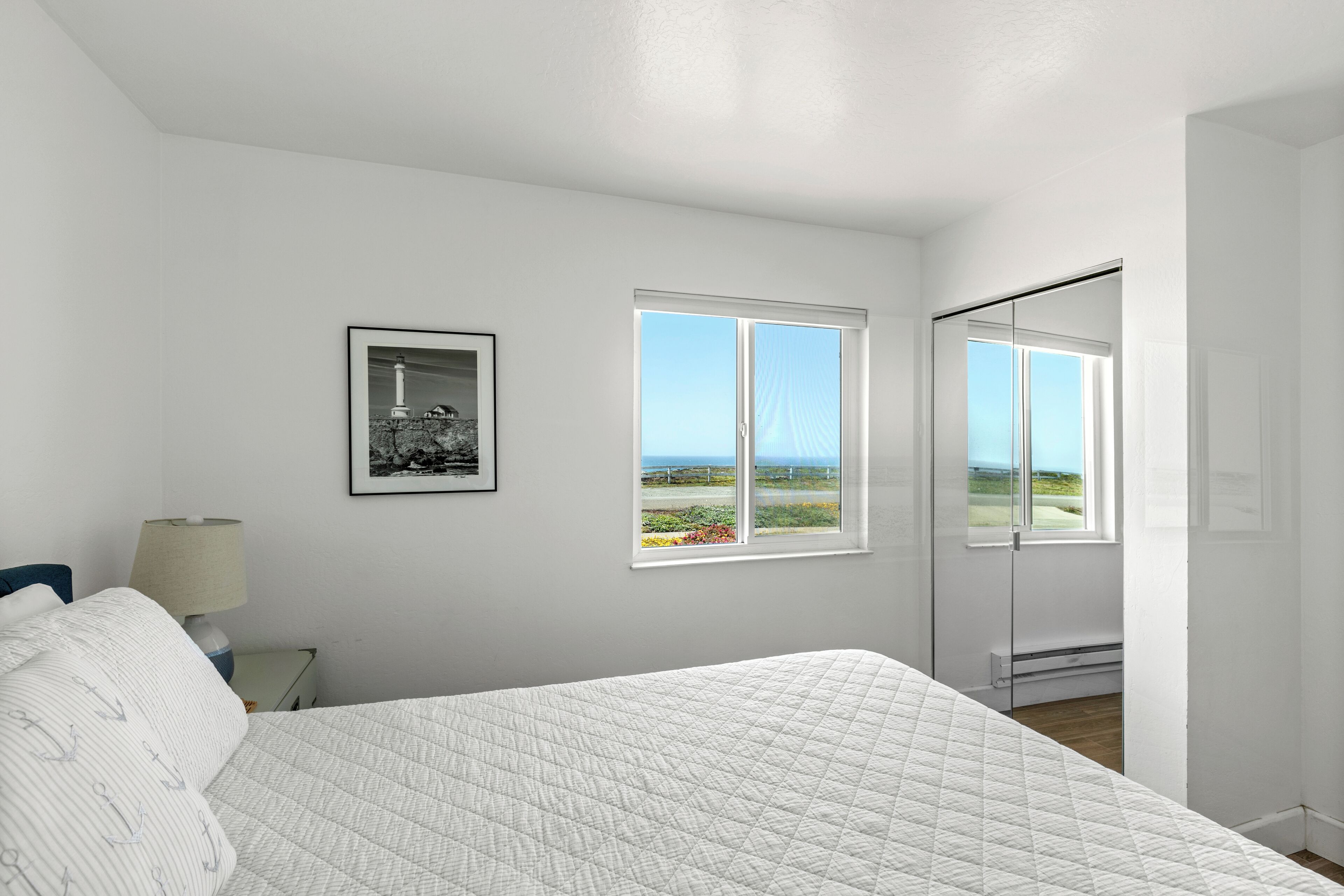 A bedroom with a bed, a nightstand, a lamp, a framed picture, and a window with a view of the outdoors.