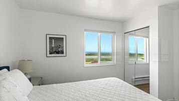 A bedroom with a bed, a nightstand, a lamp, a framed picture, and a window with a view of the outdoors.