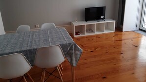 A room with a dining table, four chairs, a television on a white stand, and a large window.