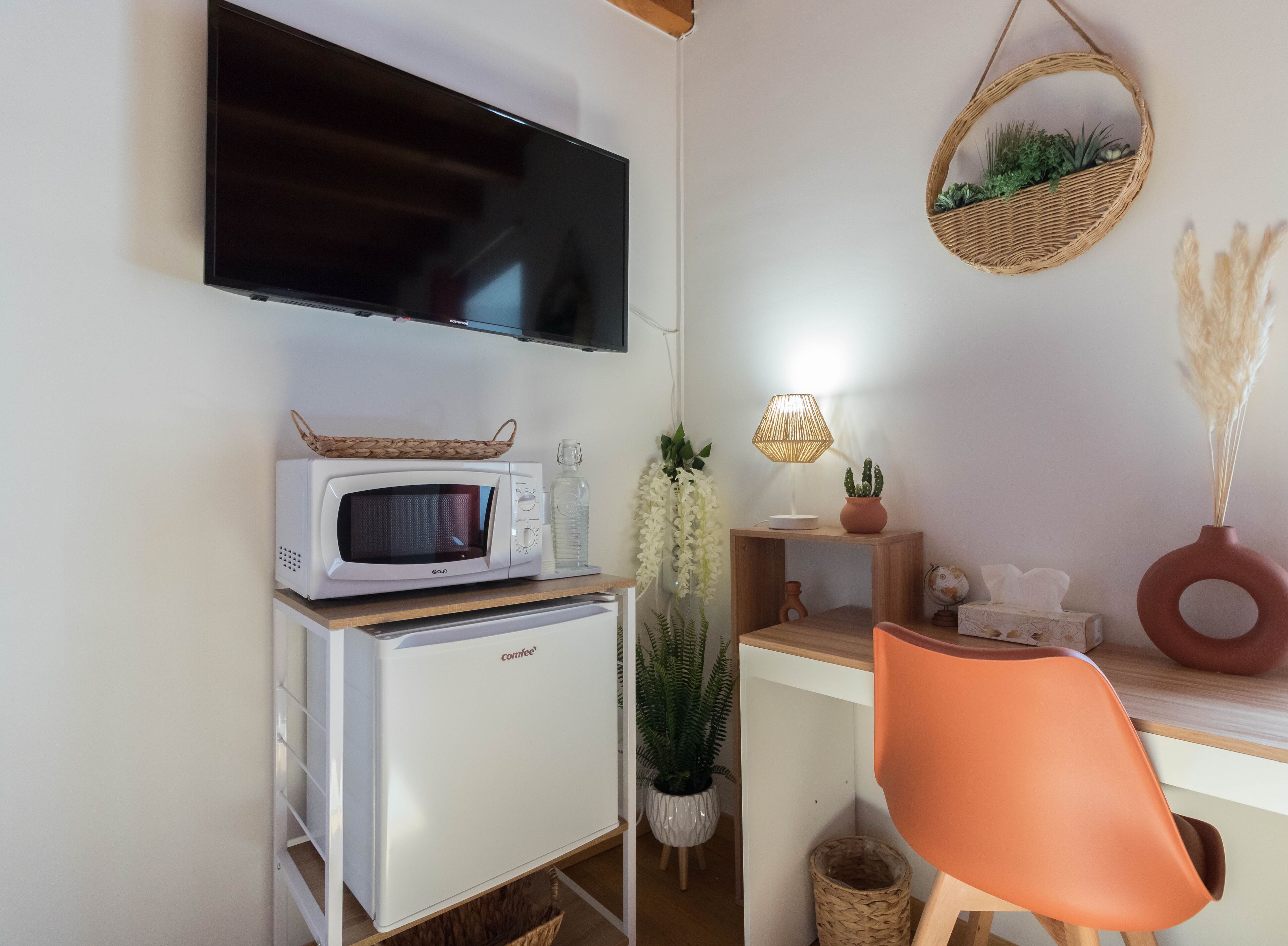 Ett litet, praktiskt kök med mikrovågsugn, ett litet kylskåp, en tv som är monterad på väggen, en korg av rotting med växter, en lampa och en stol.
