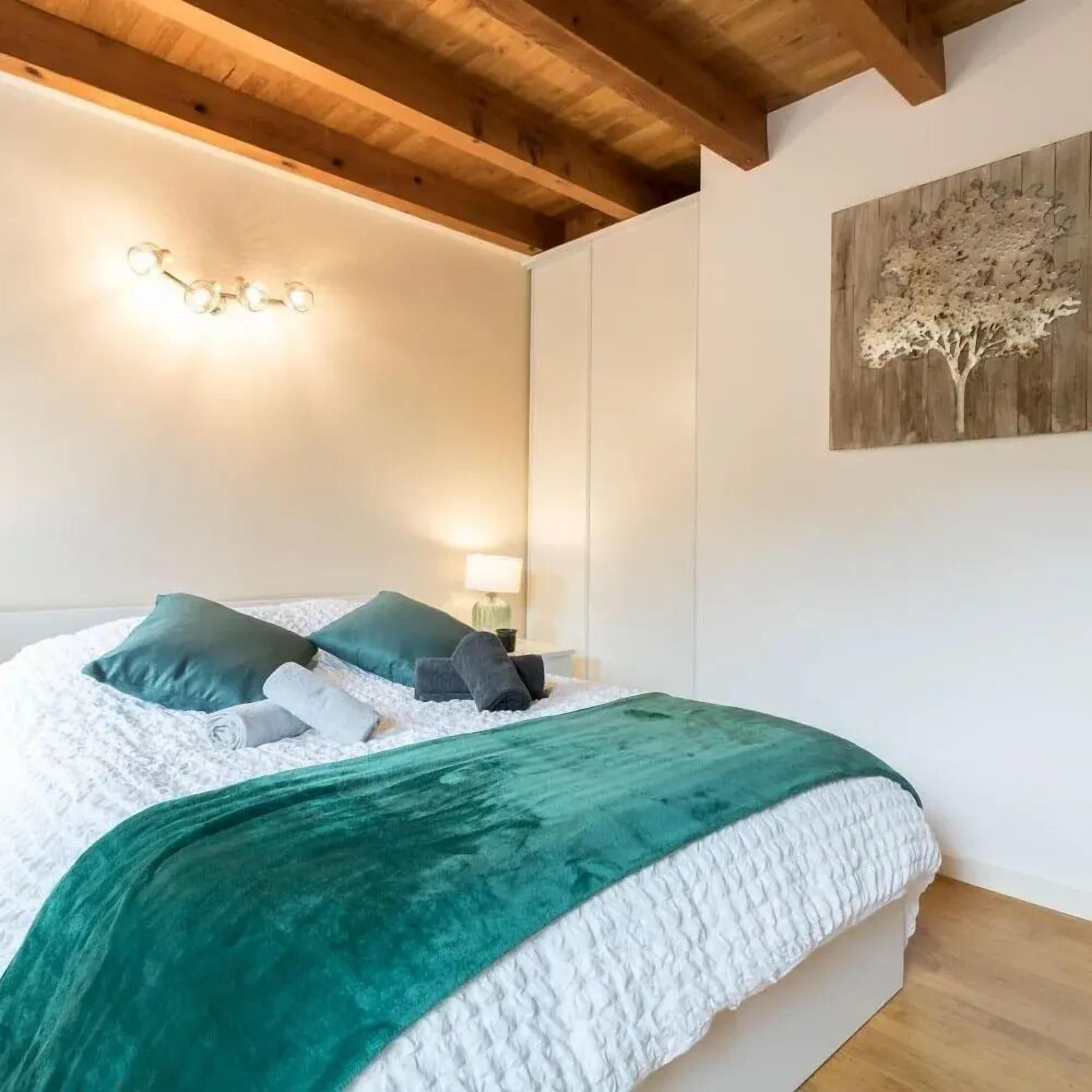 Un letto rifatto con cura, con copripiumino bianco e azzurro, due cuscini azzurri e una coperta azzurra. Sopra il letto, una lampada a parete e, sulla parete adiacente, un'opera d'arte raffigurante un albero di legno.