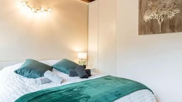 Un letto rifatto con cura, con copripiumino bianco e azzurro, due cuscini azzurri e una coperta azzurra. Sopra il letto, una lampada a parete e, sulla parete adiacente, un'opera d'arte raffigurante un albero di legno.