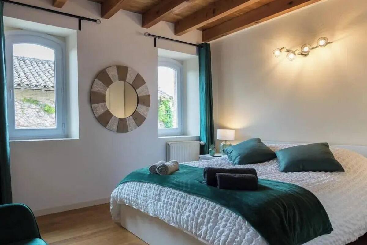 A bedroom with a bed, two windows, a mirror, and a wall-mounted light fixture.
