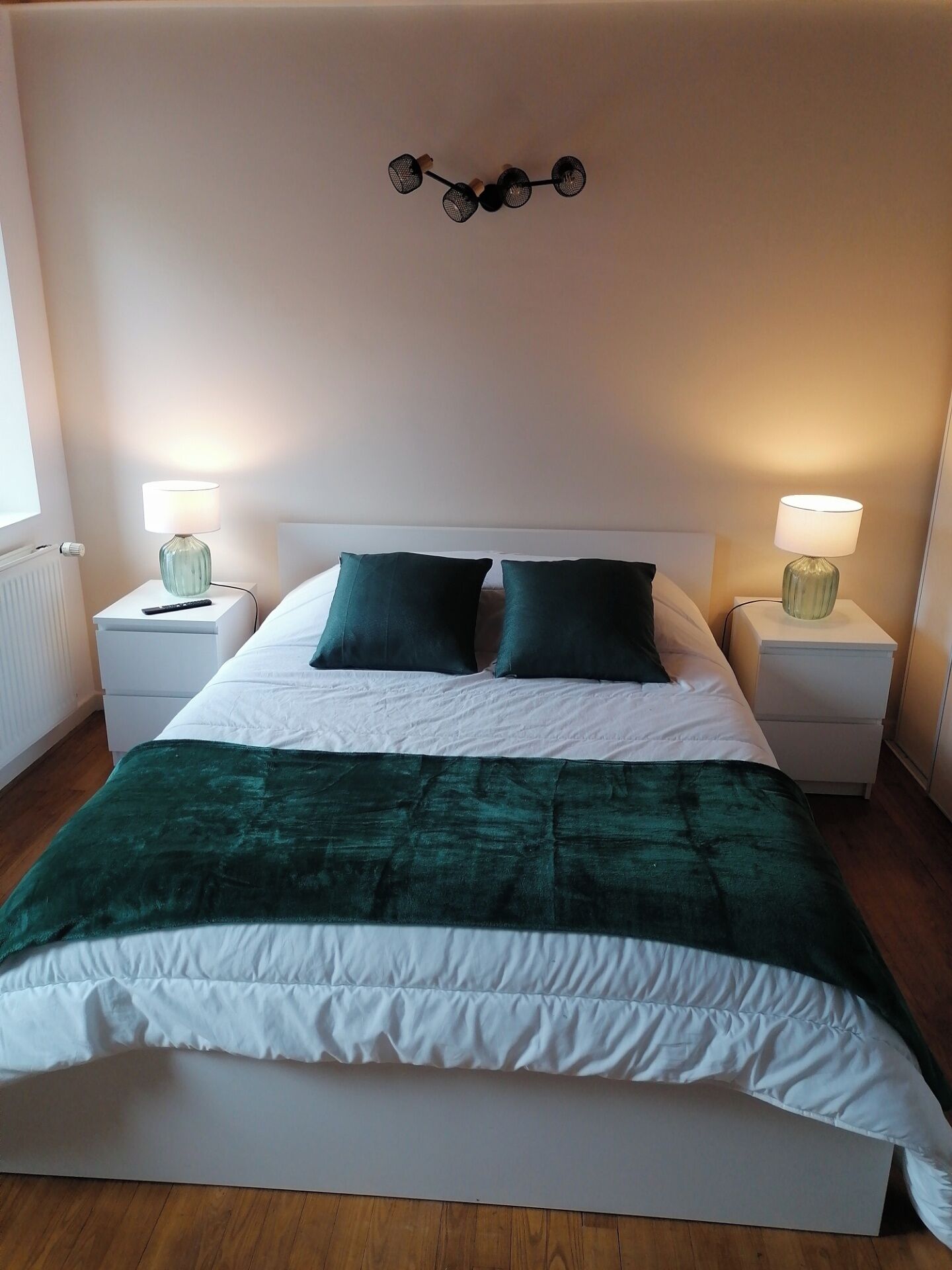 Een netjes opgemaakt bed met een groene deken, twee witte kussens en een hoofdeinde met een decoratieve lamp. Er zijn twee nachtkastjes met lampen en een raam aan de zijkant.