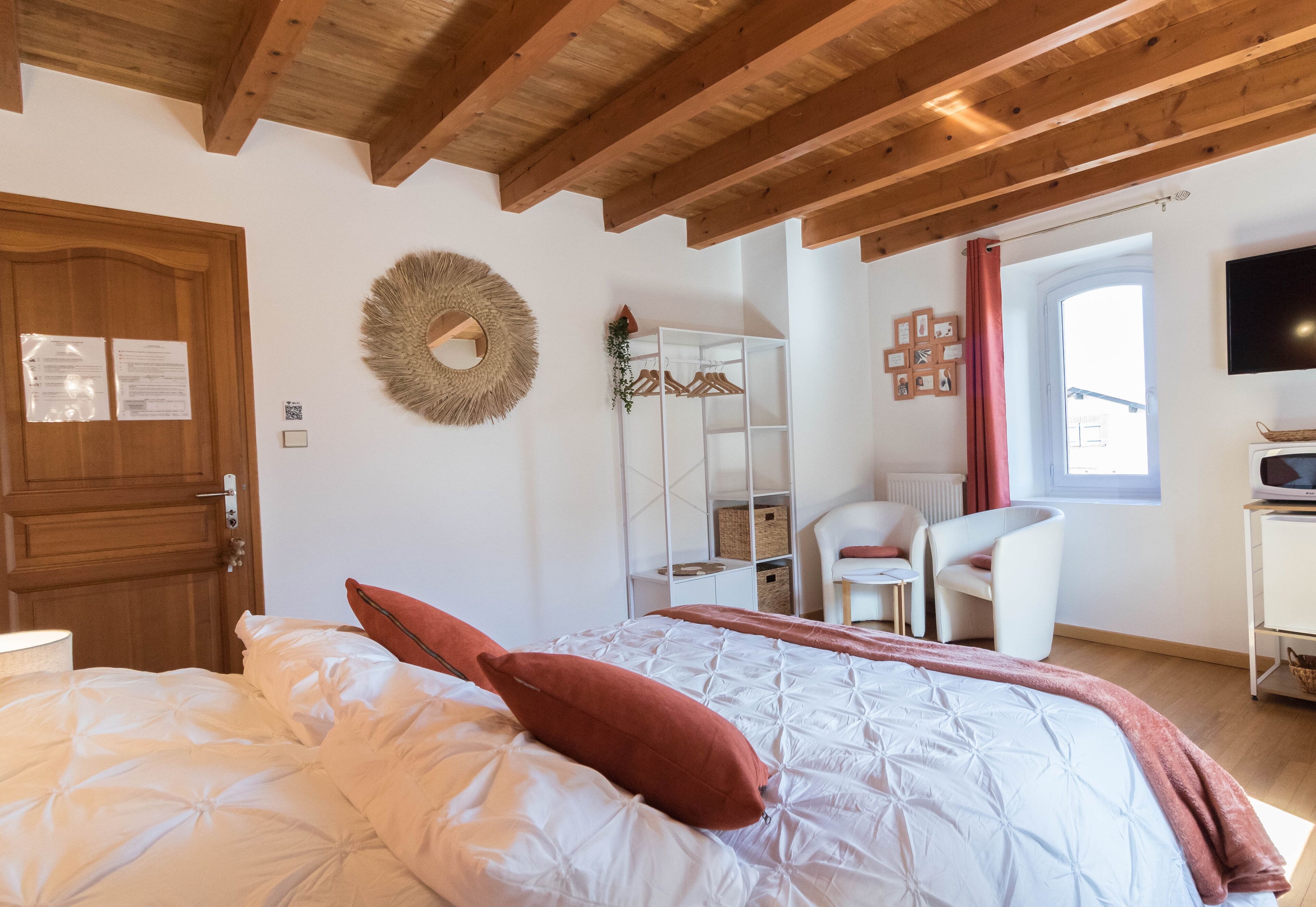 A bedroom with a wooden ceiling, a bed with white bedding and a red pillow, a white wardrobe, a mirror, a chair, and a TV on a stand.
