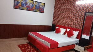 A room with a bed, a red and white bedspread, a red headboard, a red and white pillow set, a red patterned rug, a wooden nightstand, a mirror, and a wall with a geometric pattern. - Avexia Fun City Resort  (Mirzapur)