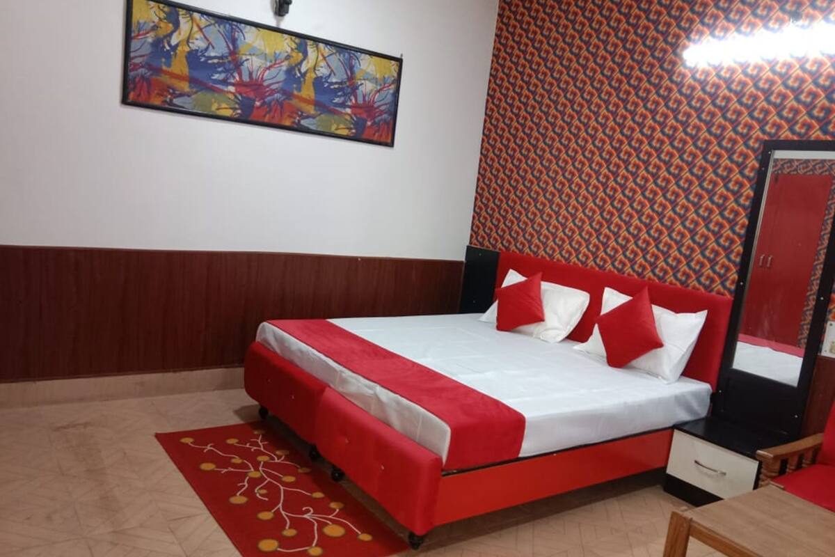 Una habitación con una cama, una colcha rojo y blanco, un cabecero rojo, un juego de almohadas rojo y blanco, una alfombra con diseño rojo, una mesita de noche de madera, un espejo y una pared con un patrón geométrico.