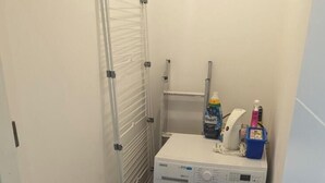 Une petite buanderie comprenant un lave-linge, un étendoir et un petit coffre de rangement servant de repose-pied.