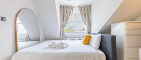A modern bedroom with a bed, a bedside table, a mirror, and a window with curtains.