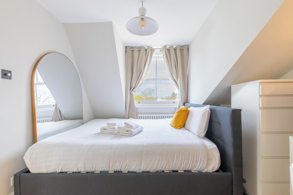 A modern bedroom with a bed, a bedside table, a mirror, and a window with curtains.