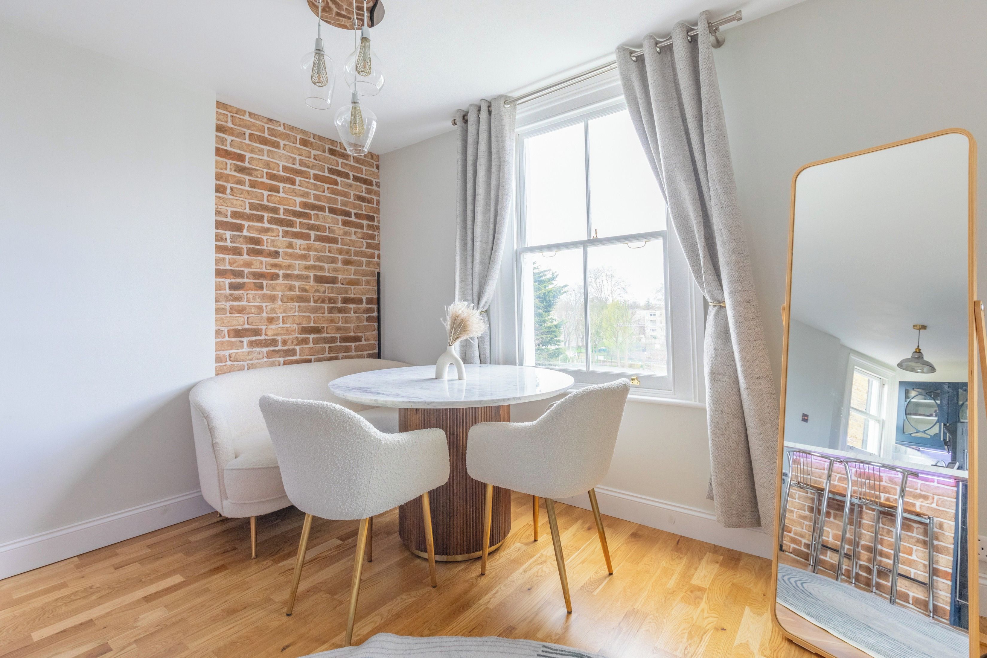 A dining area with a brick wall, a round table, two chairs, and a view through a window with curtains.