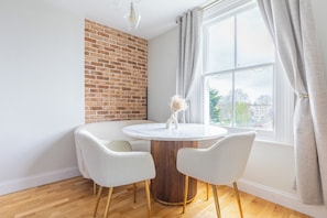 A dining area with a round table, two chairs, a brick wall, and a window with curtains. - 2BDR Hidden Gem in the Heart of Clapham (London)