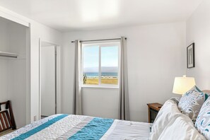 A bedroom with a bed, a window with a view of the sea, a bedside table with a lamp, and a door leading to another room. - Point Arena Lighthouse - Assistant Keeper's House 2 (Point Arena)