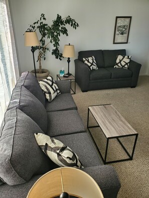 Ein Wohnzimmer mit einem Ecksofa, zwei Beistelltischen, einer Stehlampe, einer Topfpflanze und einem gerahmten Bild an der Wand.