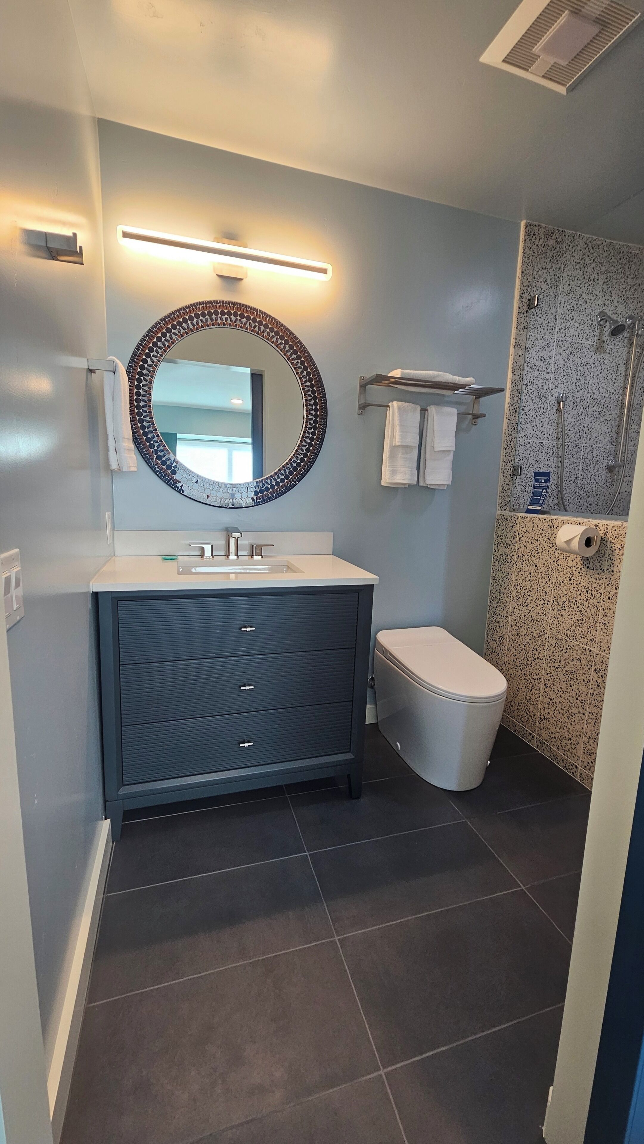 Une salle de bain moderne avec un meuble-lavabo aux tons foncés, un miroir rond doté d’un cadre en mosaïque, des toilettes et un espace douche avec un mur recouvert de carreaux à motifs.