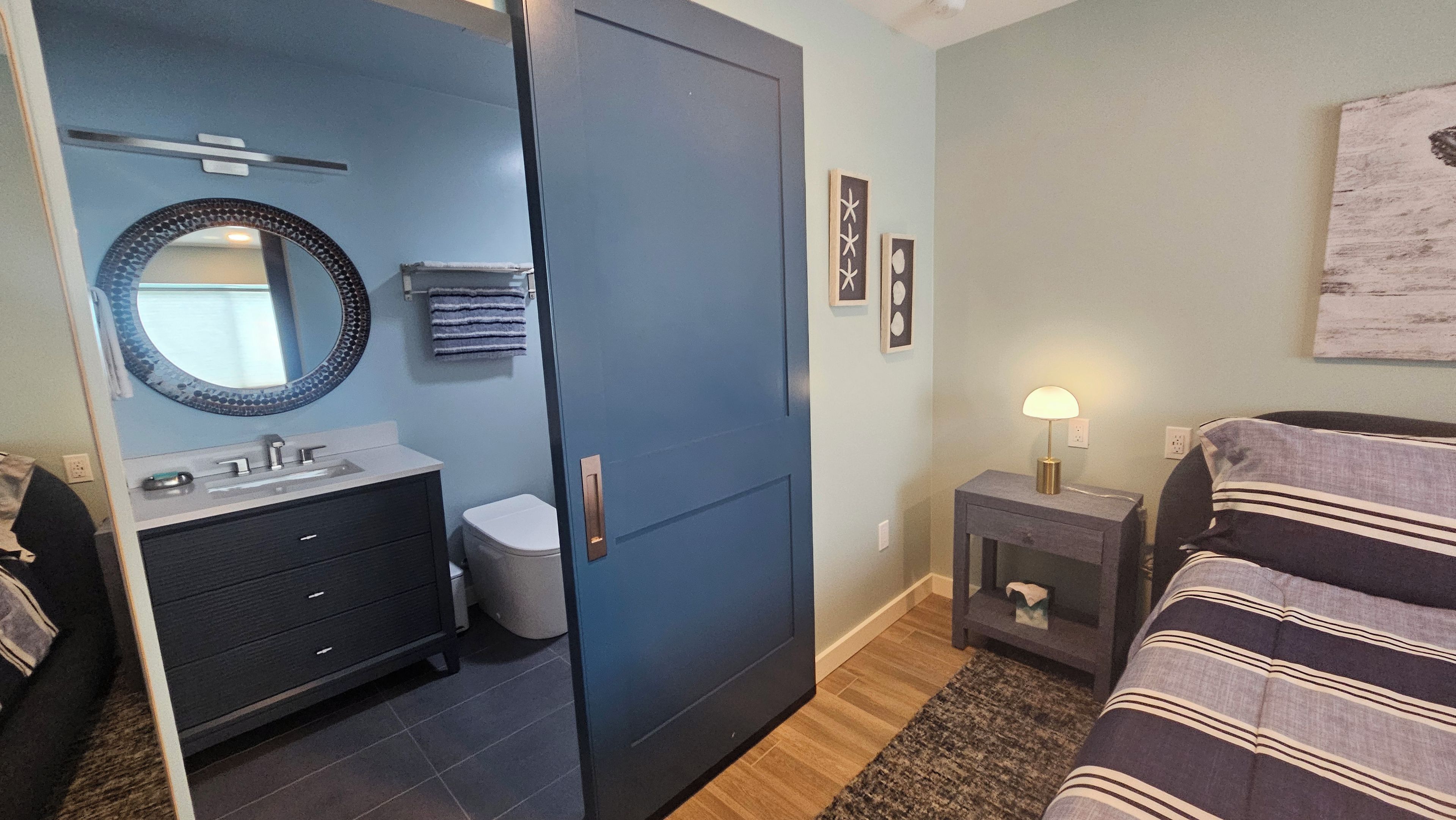 A bedroom with a bed, a bedside table with a lamp, a wardrobe, a bathroom with a sink and mirror, and a small rug.