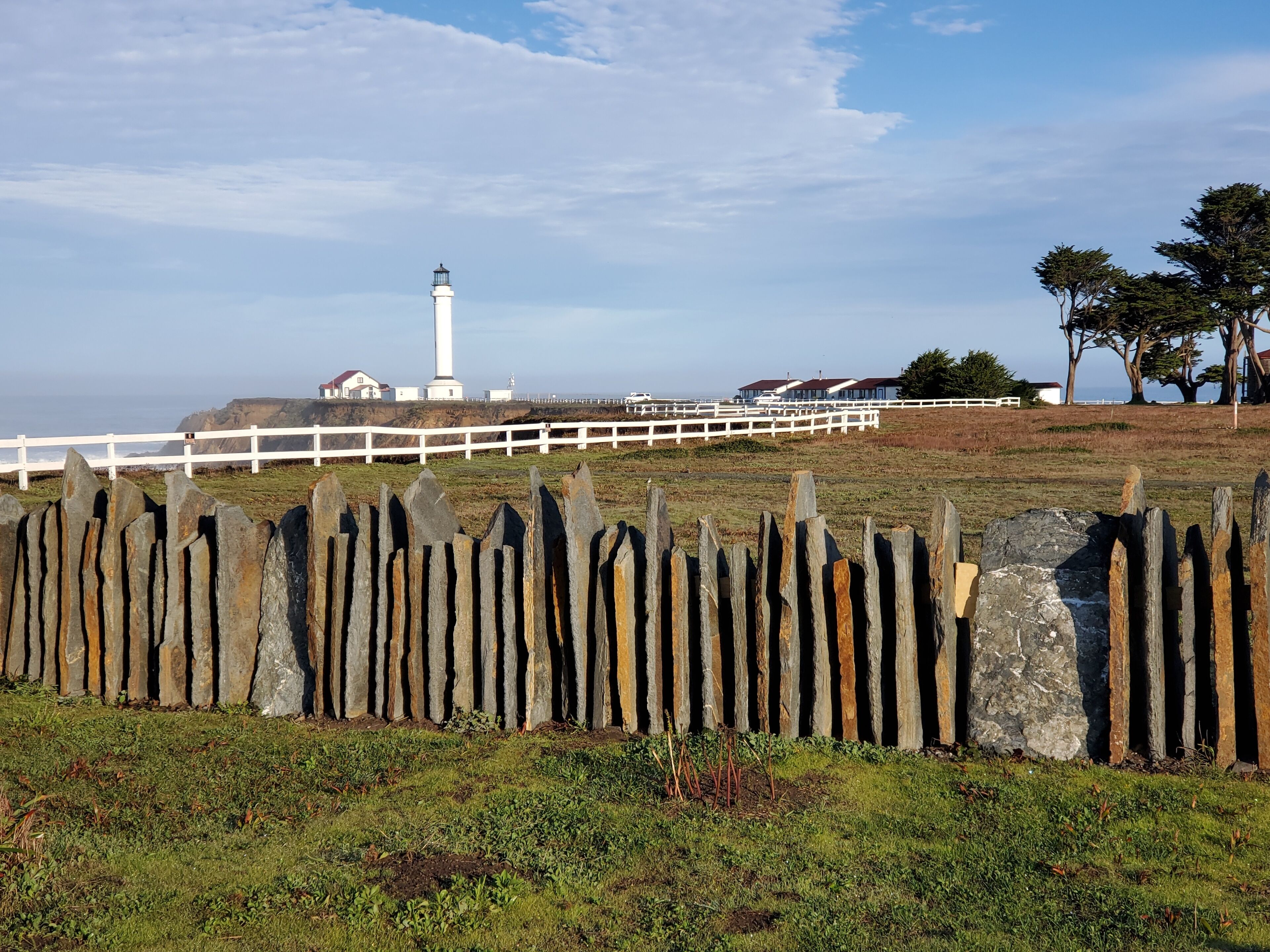 A coastal landscape with a lighthouse, white fence, and houses in the background.