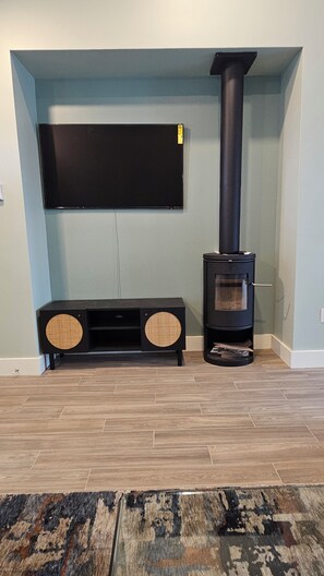 A living room with a flat-screen TV mounted on the wall, a black wood-burning stove, and a black entertainment unit with two speakers.
