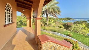 A covered outdoor area with a stone bench, a tiled floor, and a view of a coastal landscape with palm trees and a bridge.