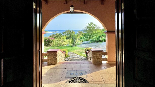 An open doorway leading to a patio with a stone bench, a view of the sea, and a clear sky.