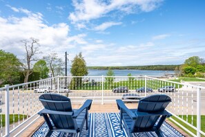 A deck with two Adirondack chairs overlooking a body of water, a road with cars, and a grassy area with trees. - The Deckhouse | Water Views, Walk Downtown, Sauna (Fish Creek)