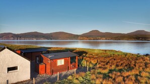 A small building with a red exterior, situated near a body of water with hills in the background. - Harlosh Log Cabins, Cabin No 2 (Dunvegan)