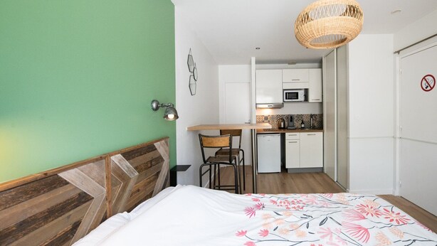 A bedroom with a wooden headboard, a bed with a floral bedspread, a dining area with a table and chairs, a kitchenette with a microwave, and a hanging wicker light fixture. - Central Part-Dieu - Apartment 1 (Lyon)