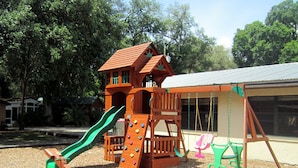 A wooden play structure with a slide and climbing wall, swings, and a small building with a roof. - Riverside Lodge Resort (Inverness)