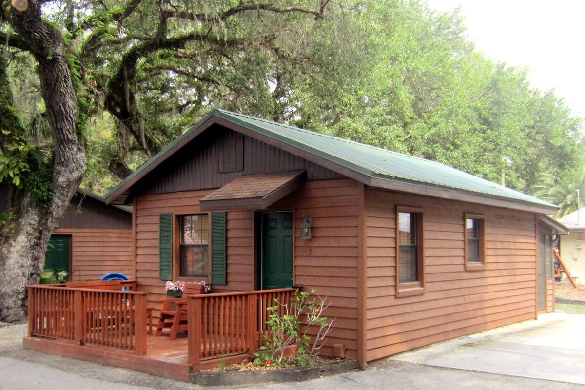 A small, single-story wooden cabin with a porch, green shutters, and a metal roof.