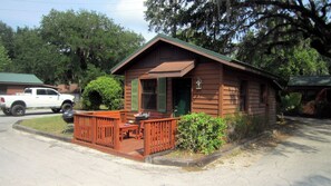 A small wooden cabin with a porch, a truck parked nearby, and surrounding greenery.