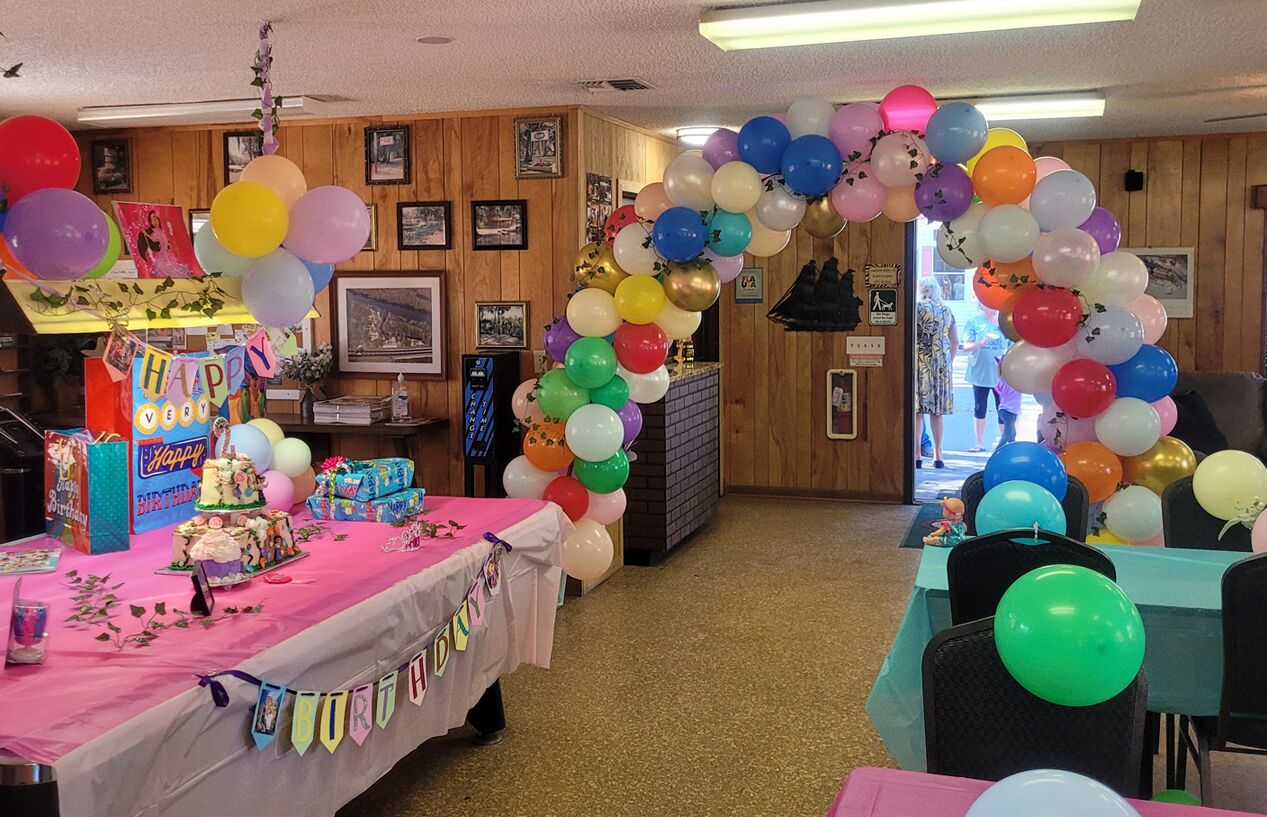 A room with a birthday celebration setup, including a table with a cake and gifts, a balloon arch, and a 'Happy Birthday' banner.