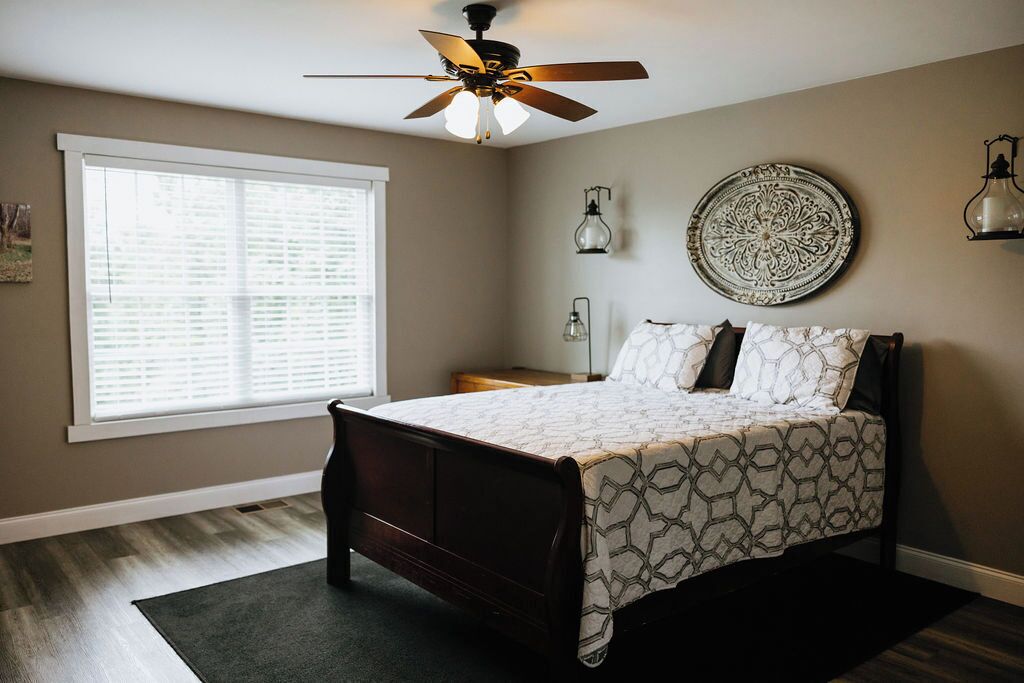 Une chambre à coucher comprenant un lit, un ventilateur de plafond, une fenêtre avec des stores, une table de chevet et des œuvres d'art décoratives sur les murs.