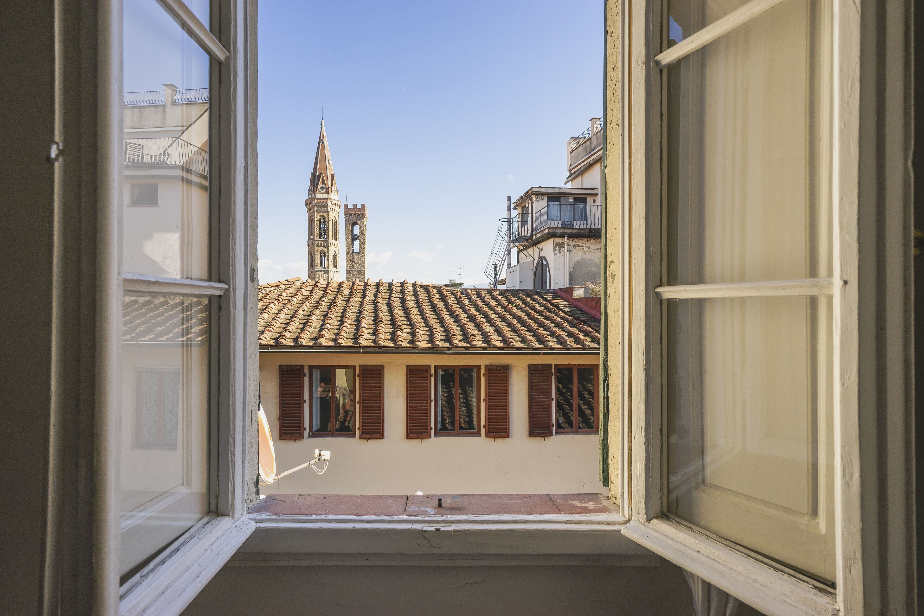Vista pela janela de um edifício com telhado de azulejos e uma torre ao fundo.