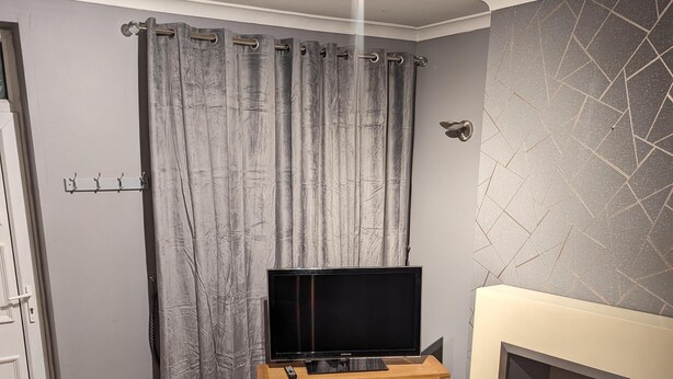 A living room with a flat-screen TV on a wooden stand, a fireplace, and a grey curtain. - 2 Bed House - Garden & Excellent Wifi (Newark)