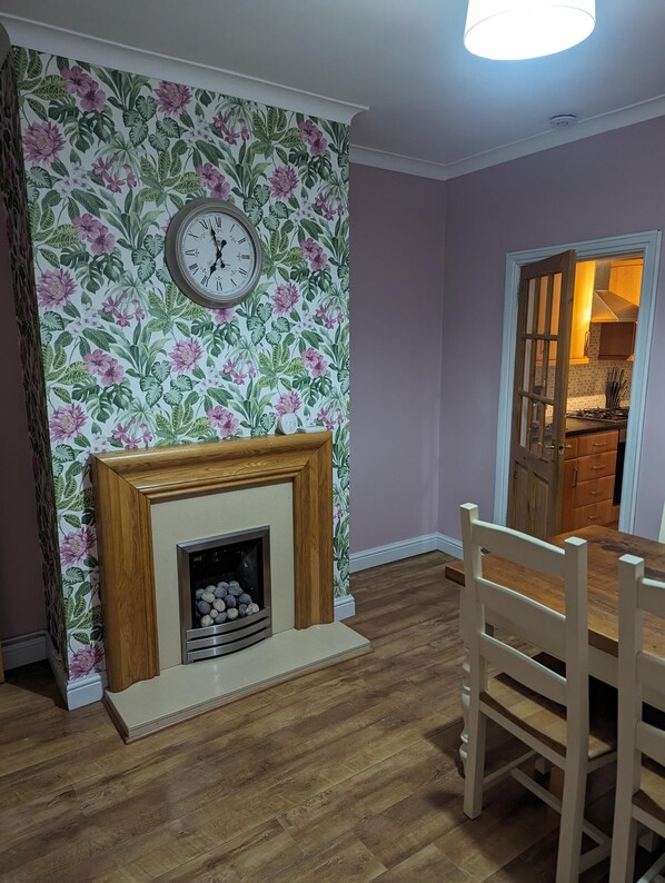 Una habitación con papel tapiz floral, una chimenea de madera, una mesa de comedor con sillas y vistas a una cocina a través de una puerta abierta.