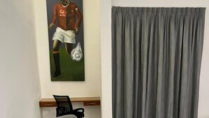Un canapé d’angle gris dans un salon moderne, avec un tableau représentant un joueur de football accroché au mur, un bureau en bois et des rideaux gris.