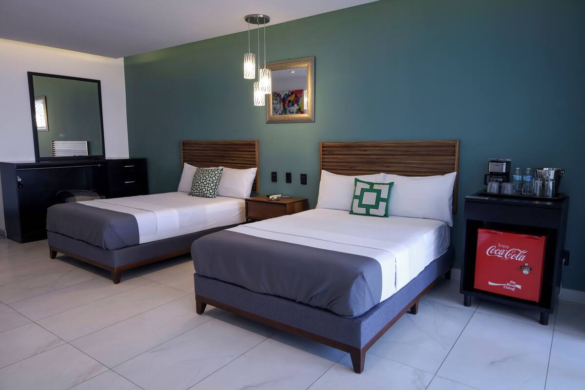 Une chambre d’hôtel avec deux lits, un bureau, un miroir, une table de chevet avec un mini-réfrigérateur contenant des canettes de Coca-Cola, et une œuvre d’art accrochée au mur.