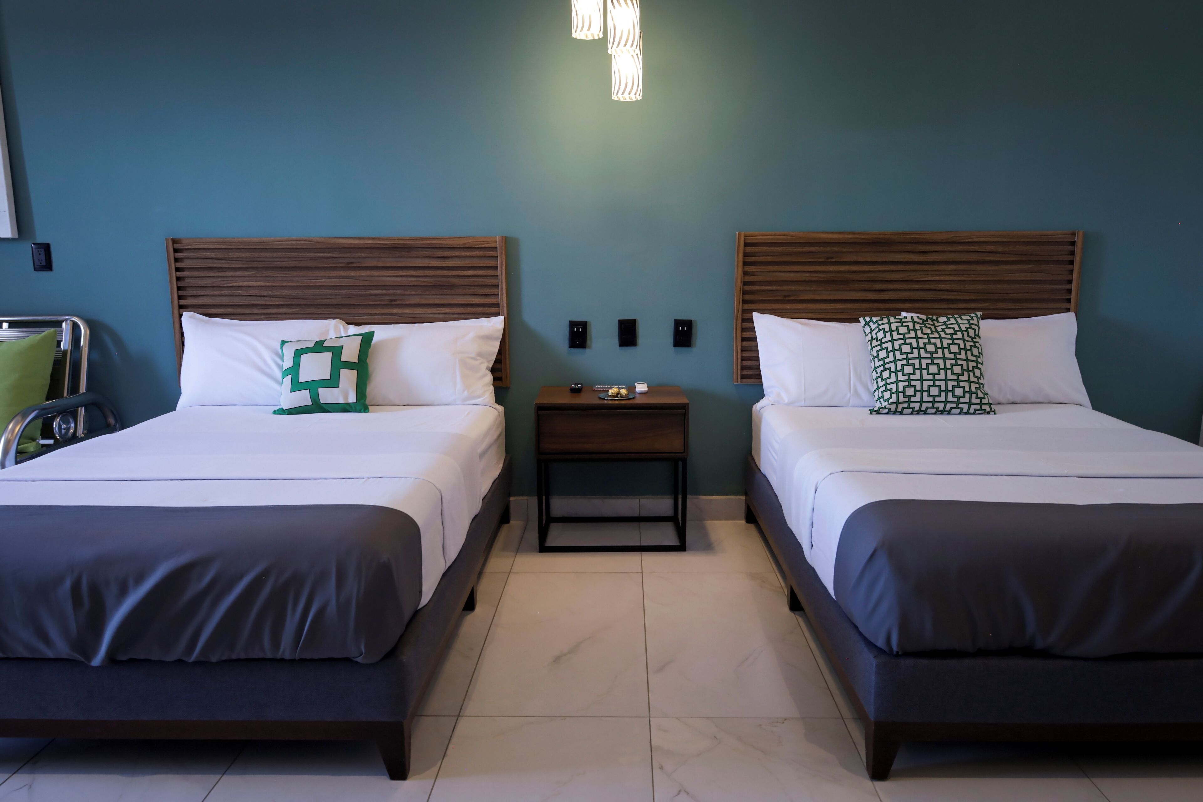 A hotel room with two beds, each with a wooden headboard, white bedding, and a small bedside table with a lamp.