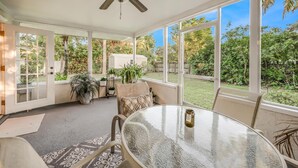 A sunroom with a ceiling fan, large windows, a round glass table, and various plants. - Naples Florida (Naples)
