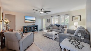 A spacious living room with a ceiling fan, a large window with blinds, a gray sofa, a white coffee table, and a bookshelf. - Naples Florida (Naples)