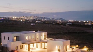 A modern two-story house with a swimming pool, surrounded by a landscaped garden and a view of a distant town at dusk.