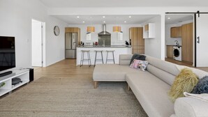 A modern living room with a large sectional sofa, a flat-screen TV, and a kitchen area with wooden cabinets and a stainless steel range hood. - House Naomi - Martinborough Holiday Home (Martinborough)