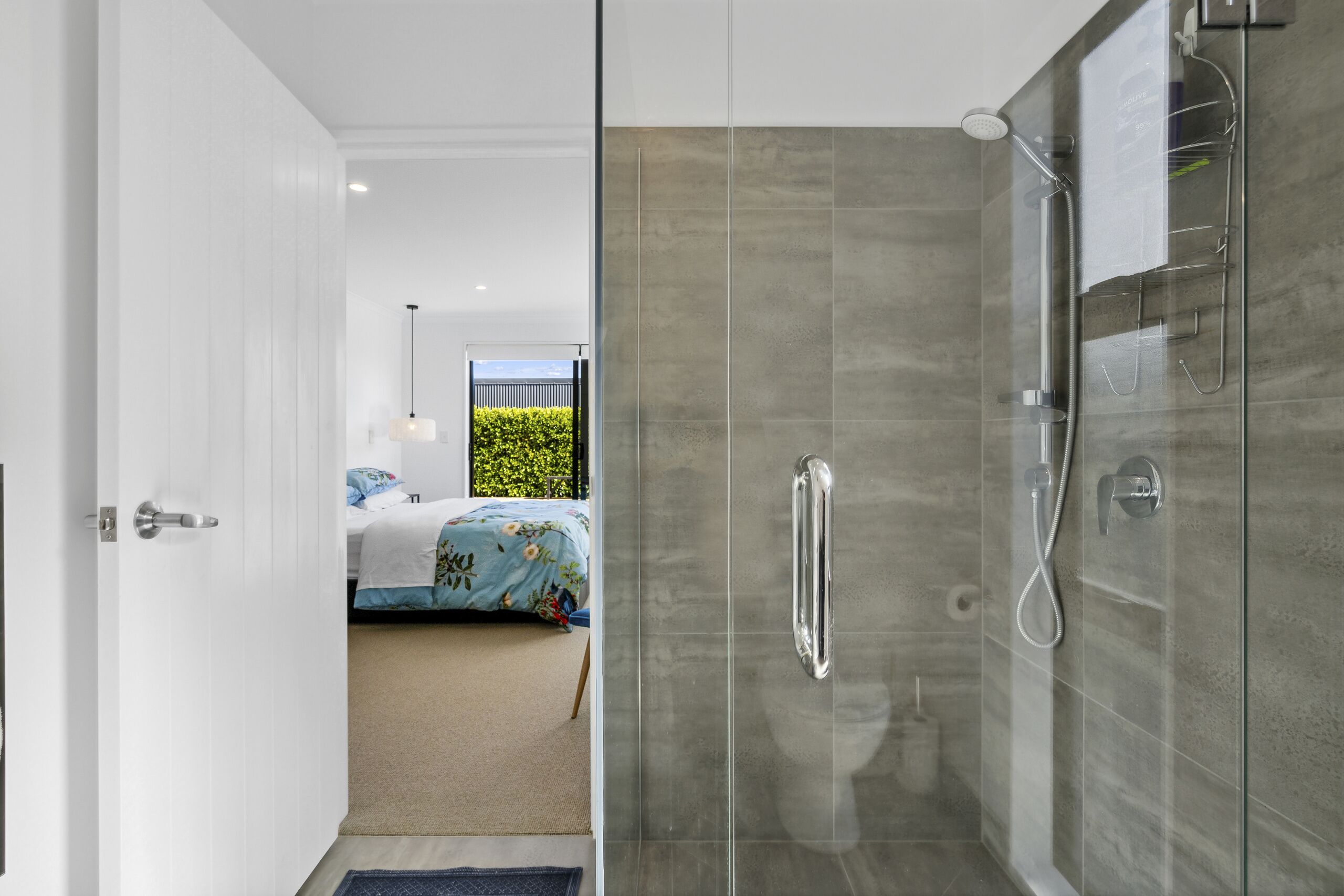 A modern bathroom with a glass shower enclosure, a visible towel rack, and a door leading to a bedroom with a bed and a view of greenery.