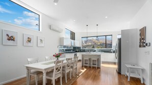 A modern kitchen with white cabinetry, a dining area with a wooden table and chairs, and a large window offering a view of a mountainous landscape. - Melmore Oasis - Cromwell Holiday Home (Cromwell)