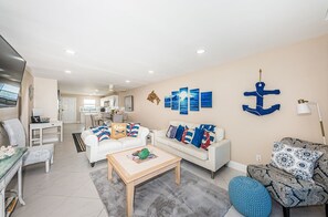 A modern living room with a white sofa, a wooden coffee table, and a patterned armchair. There is a dining area in the background with a kitchen visible. - Unobstructed Sunsets at Gulf Shores in Indian Shores 7th Floor 2/2 condo~ Gfs709 (Indian Shores)