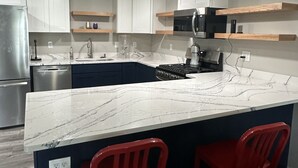 Una cocina moderna con gabinetes blancos, una isla de mármol, sillas rojas y un refrigerador de acero inoxidable.