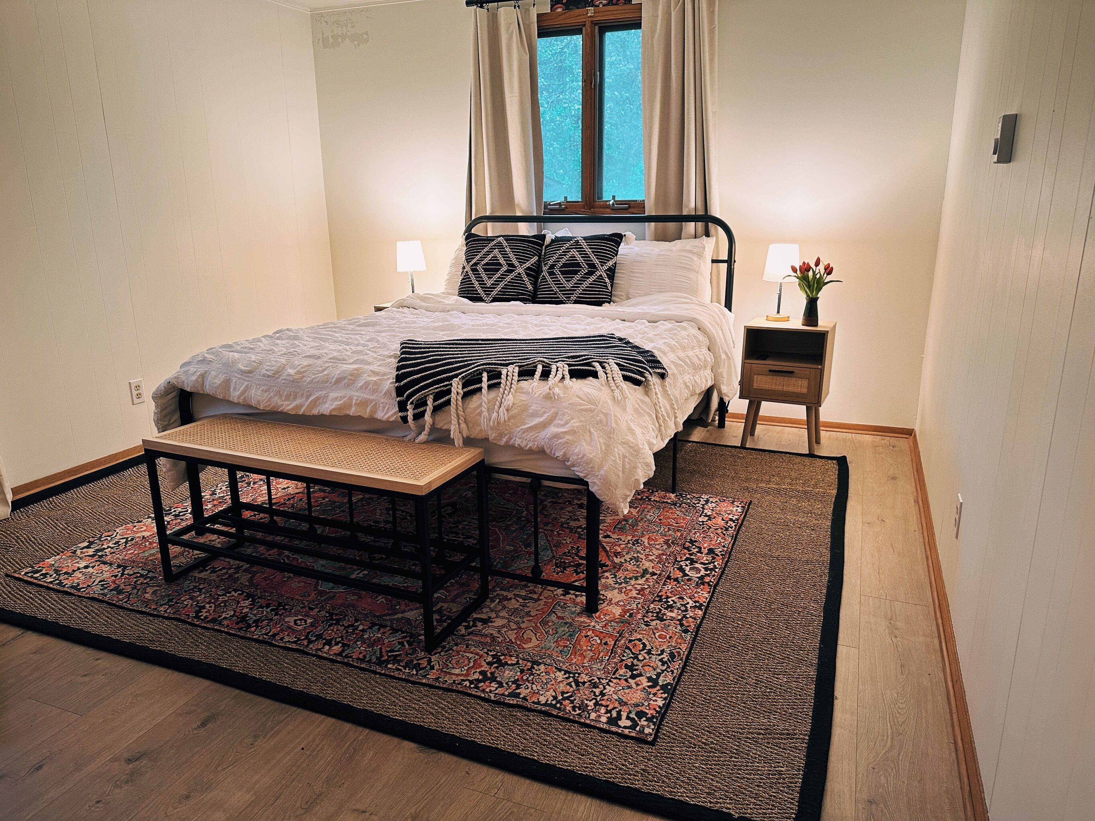 Un dormitorio ordenado con cama, mesitas de noche, un banco y una alfombra estampada.