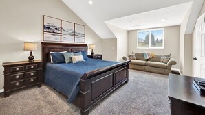 Une chambre avec un grand lit, une table de chevet, une commode, une fenêtre avec des stores et un plafond avec des poutres apparentes.